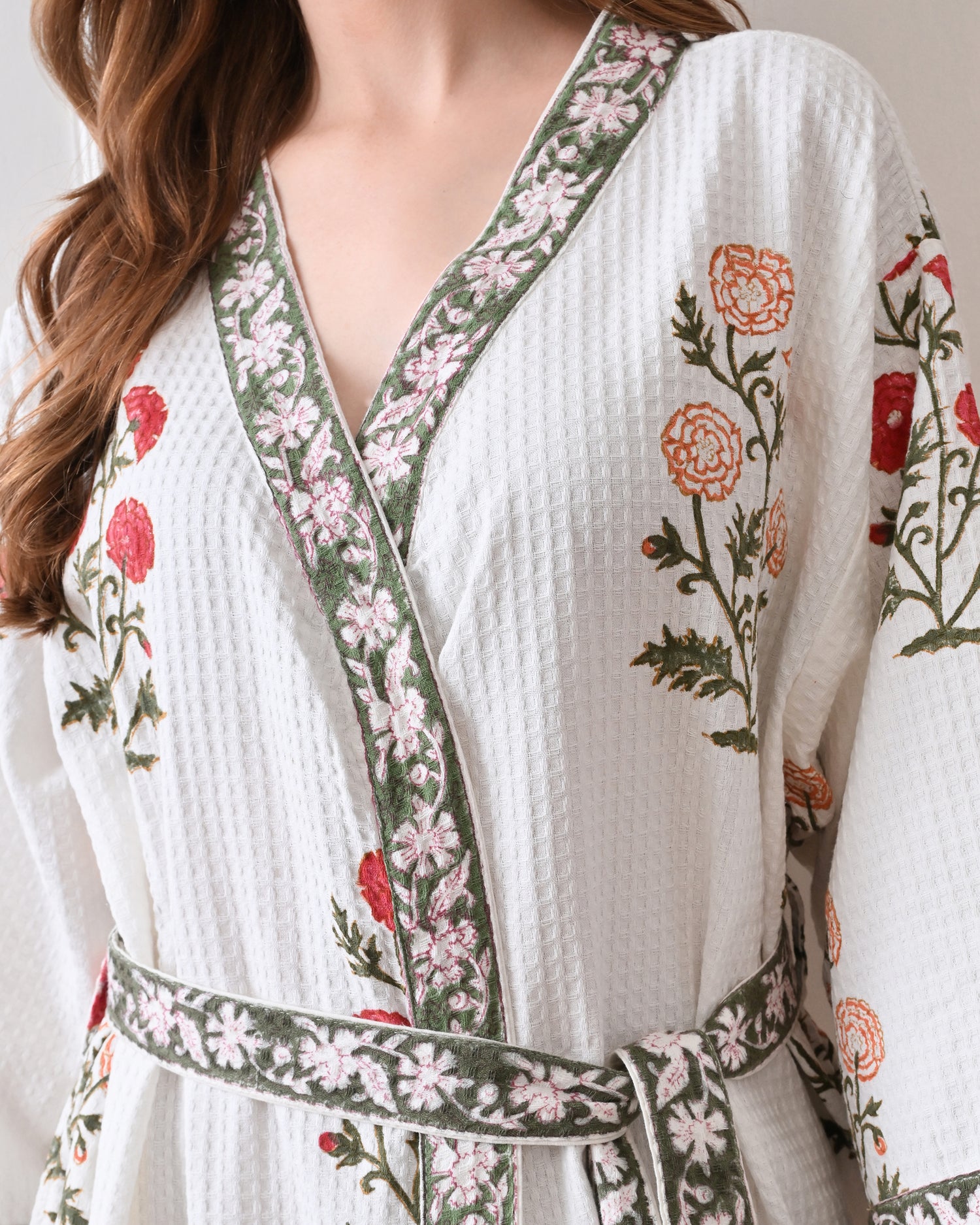 Meadow Light White Cotton Waffle Bathrobe with hand block printed floral patterns in red and orange, green leaves and green floral border, long sleeves, and waist tie belt, worn by a woman | Singhvis