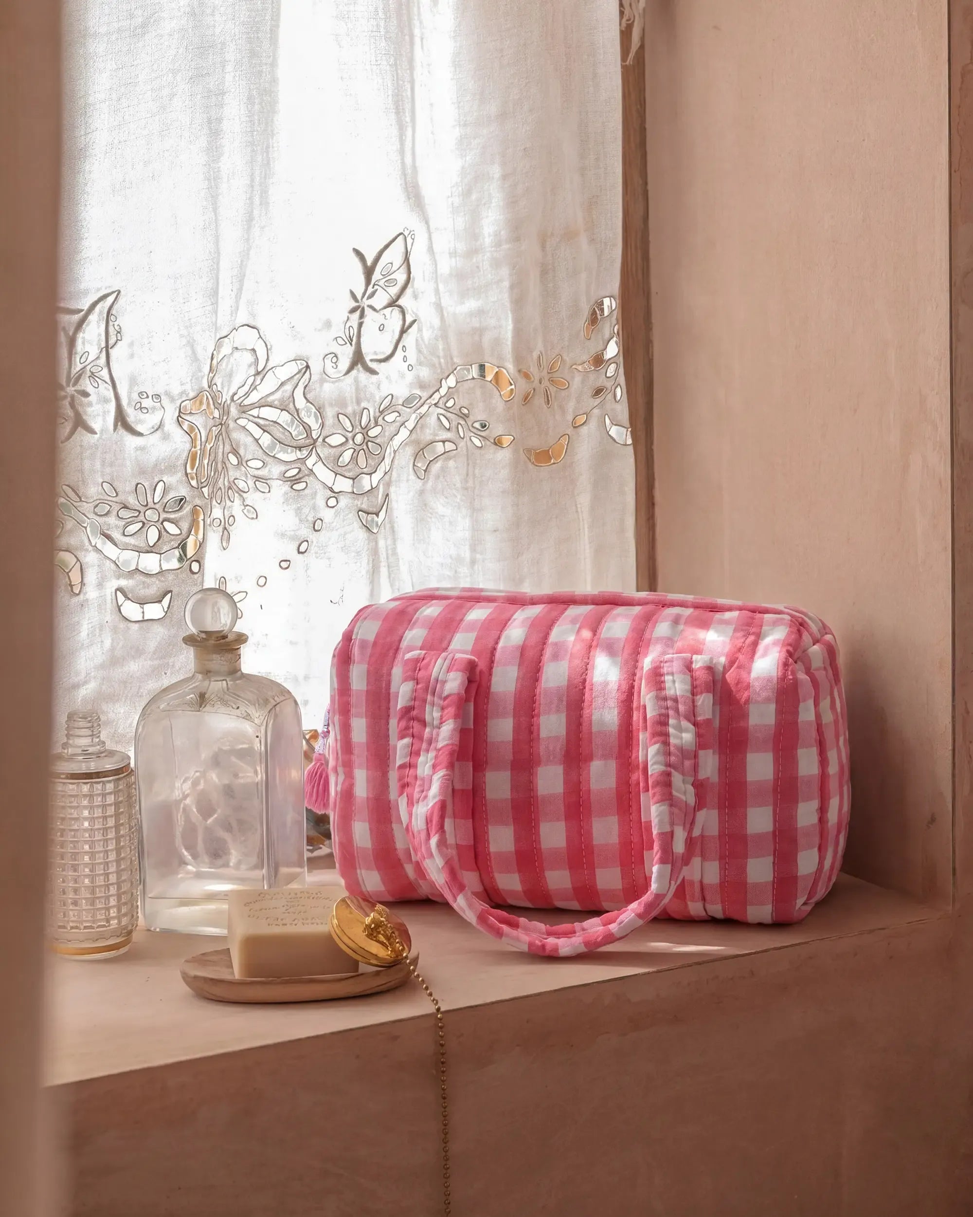 Pink gingham vanity bag with quilted texture and soft handles placed by a window next to glass bottles and a bar of soap, with a sheer curtain in the background | Singhvis