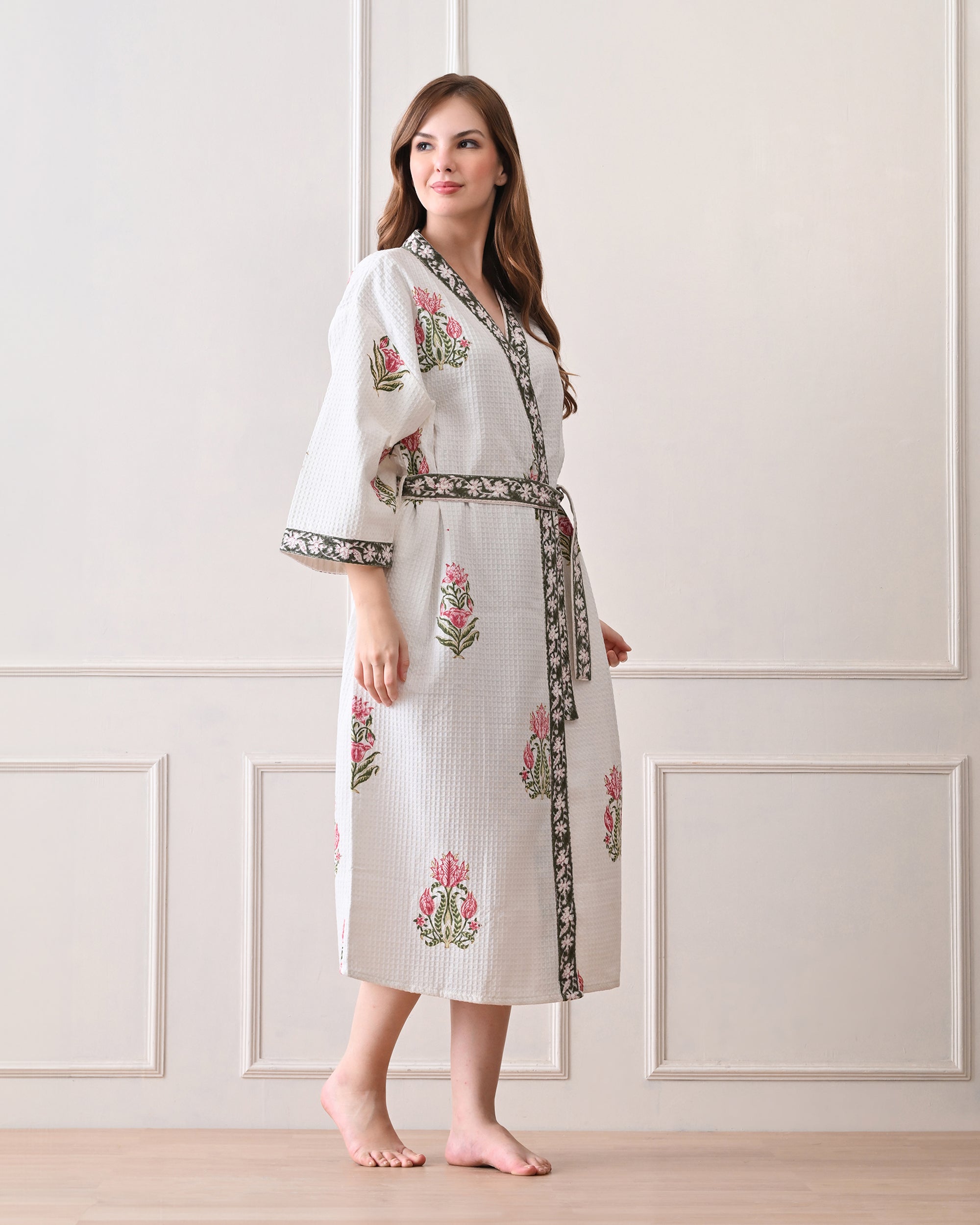 Botanic Whisper White Cotton Waffle Bathrobe with hand block printed pink tulip & lotus-inspired floral patterns, green floral border, long sleeves, and waist tie belt, worn by a woman | Singhvis