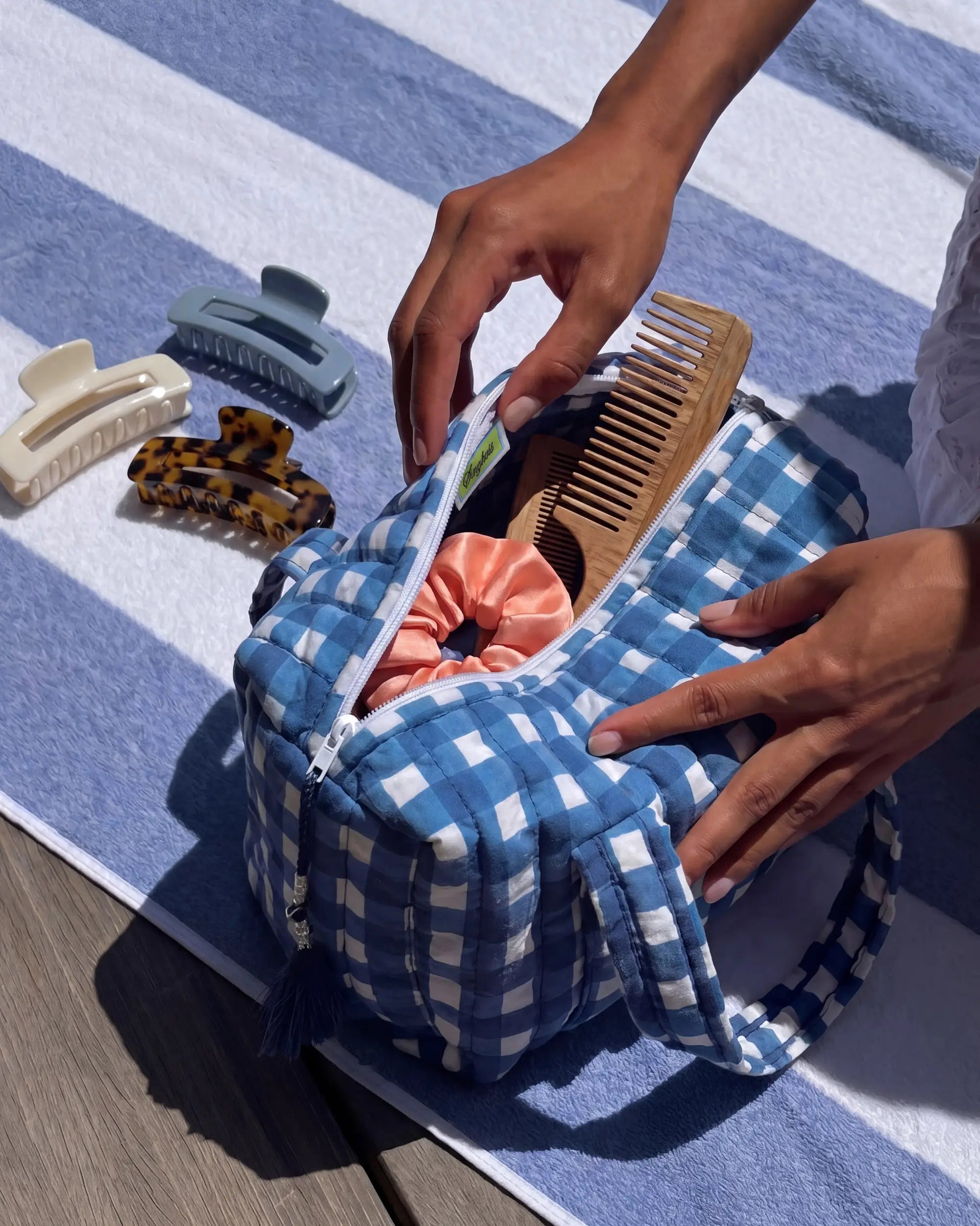 Blue gingham vanity bag with quilted texture open on a striped towel, with hands placing a comb inside and hair clips kept nearby | Singhvis