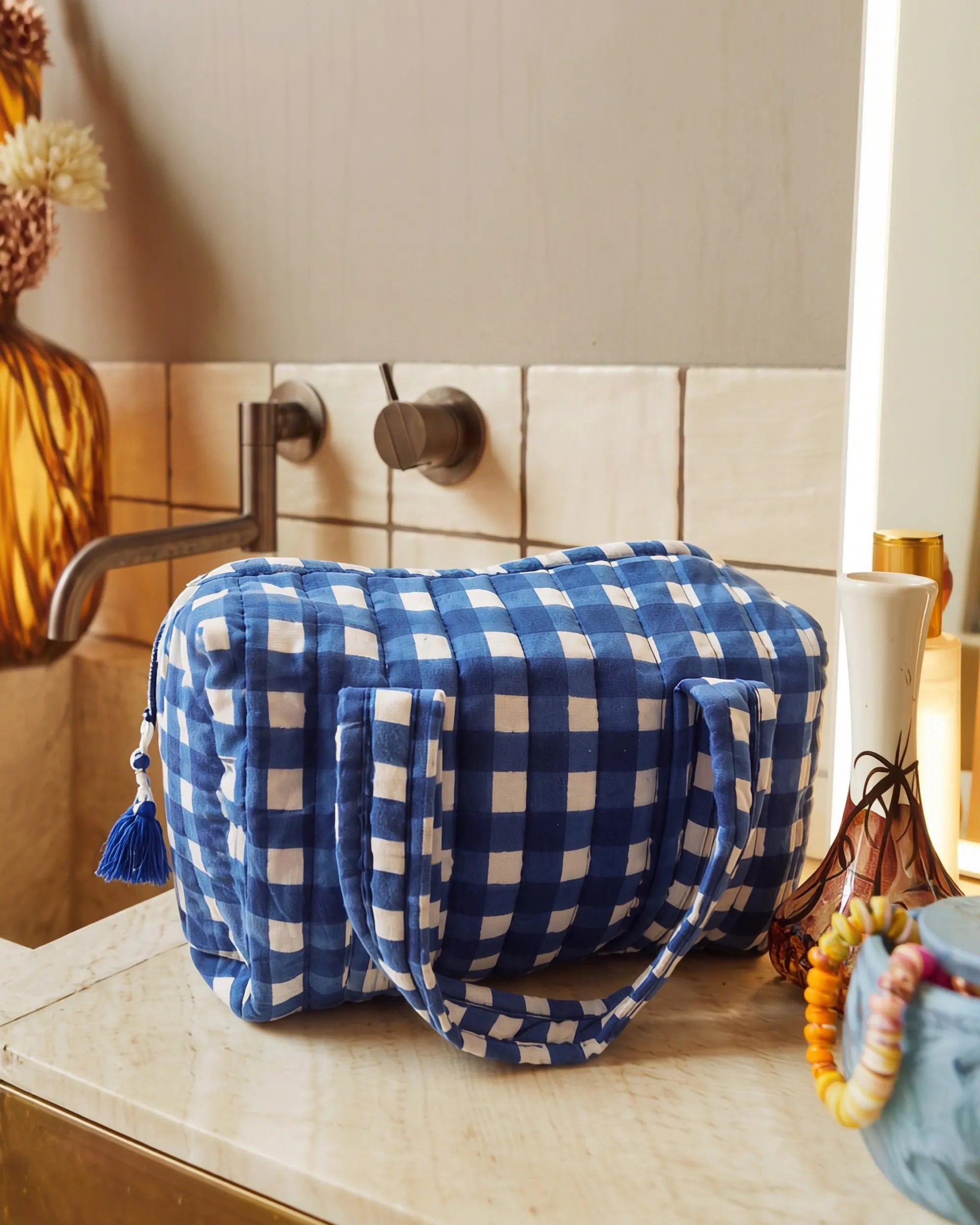 Blue gingham vanity bag with quilted texture and soft handles placed on a bathroom counter near a sink, with bottles and small items beside it | Singhvis