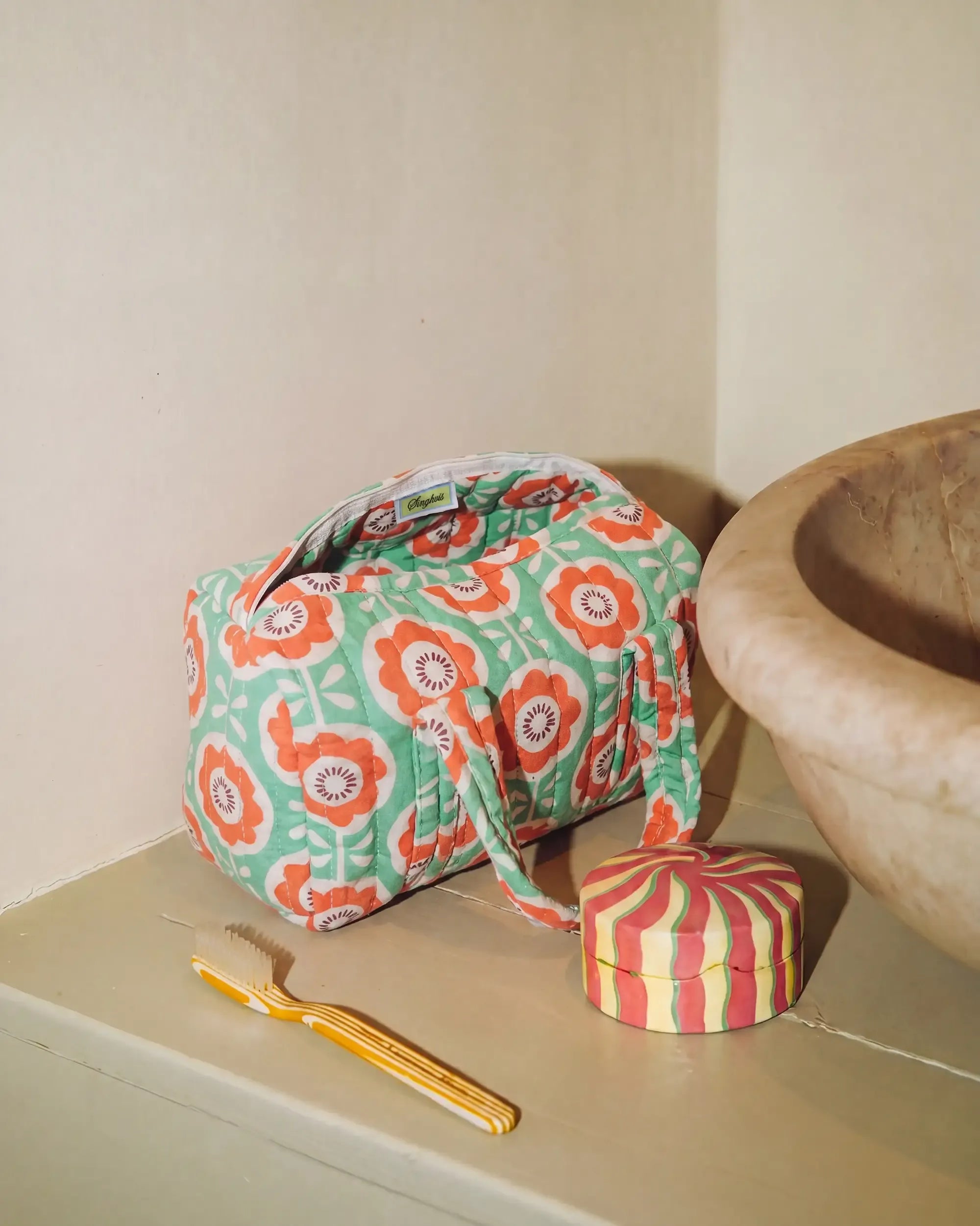 Green and orange vanity bag with floral print and quilted texture placed on a bathroom counter next to a sink, with a toothbrush and a small round box beside it | Singhvis