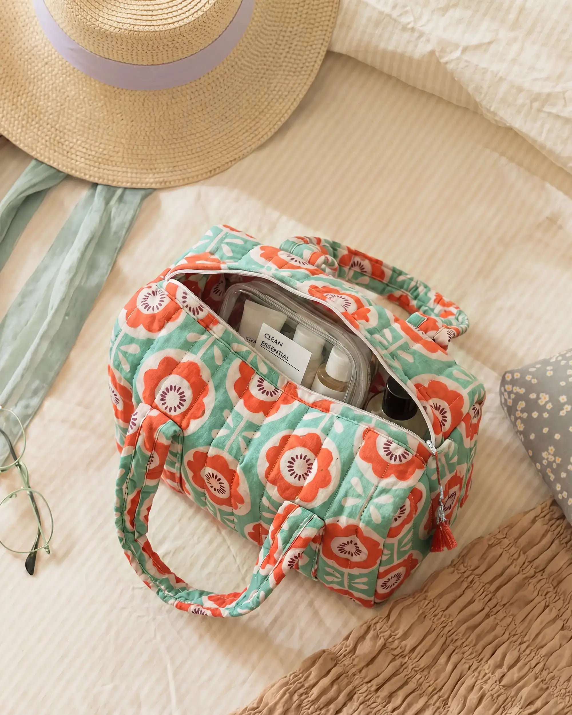 Green and orange vanity bag with floral print and quilted texture on a bed, open with bottles inside, with a hat and glasses beside it | Singhvis