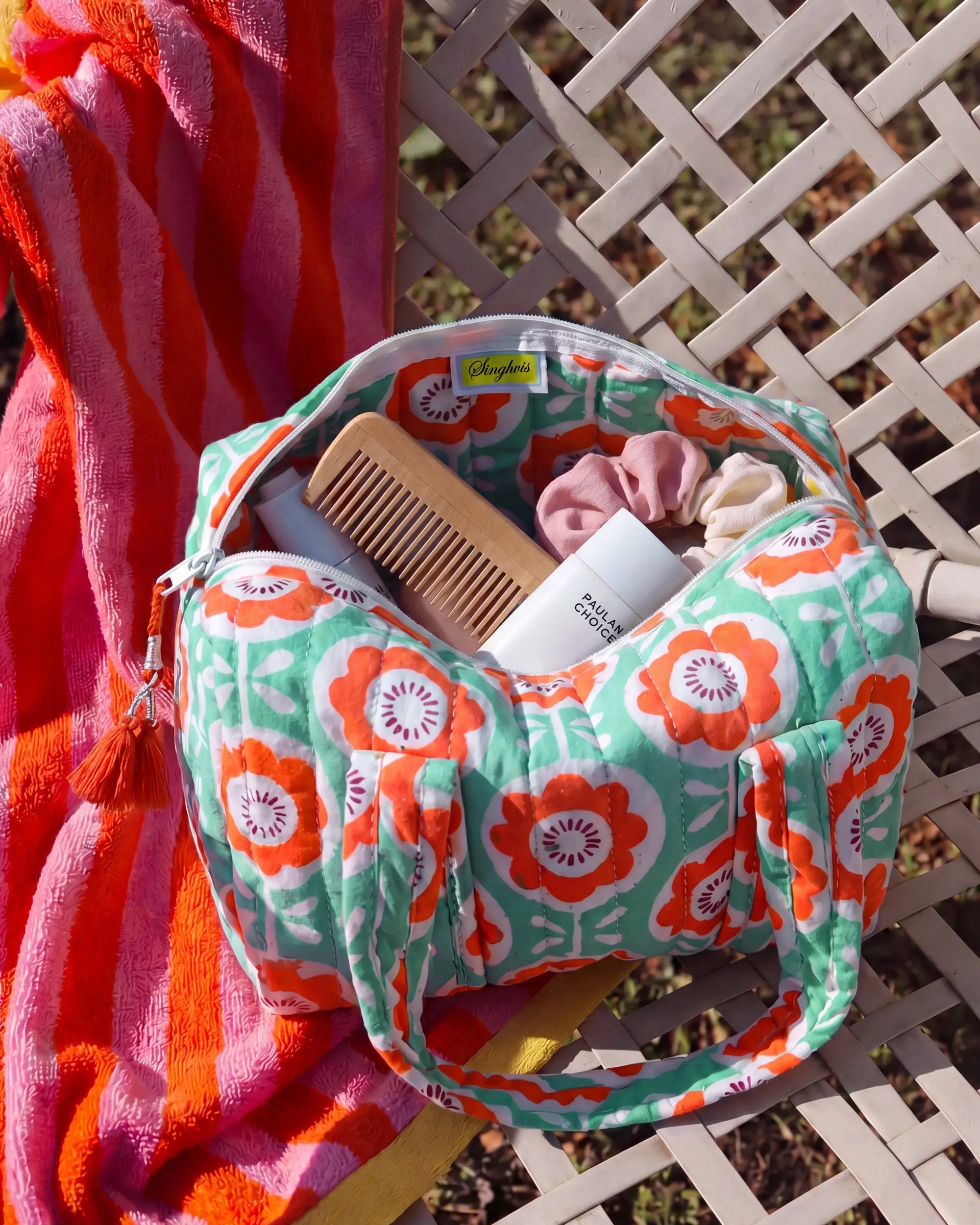 Green and orange vanity bag with floral print and quilted texture on a chair, open with a comb, scrunchies, and bottles inside, with a towel next to it | Singhvis