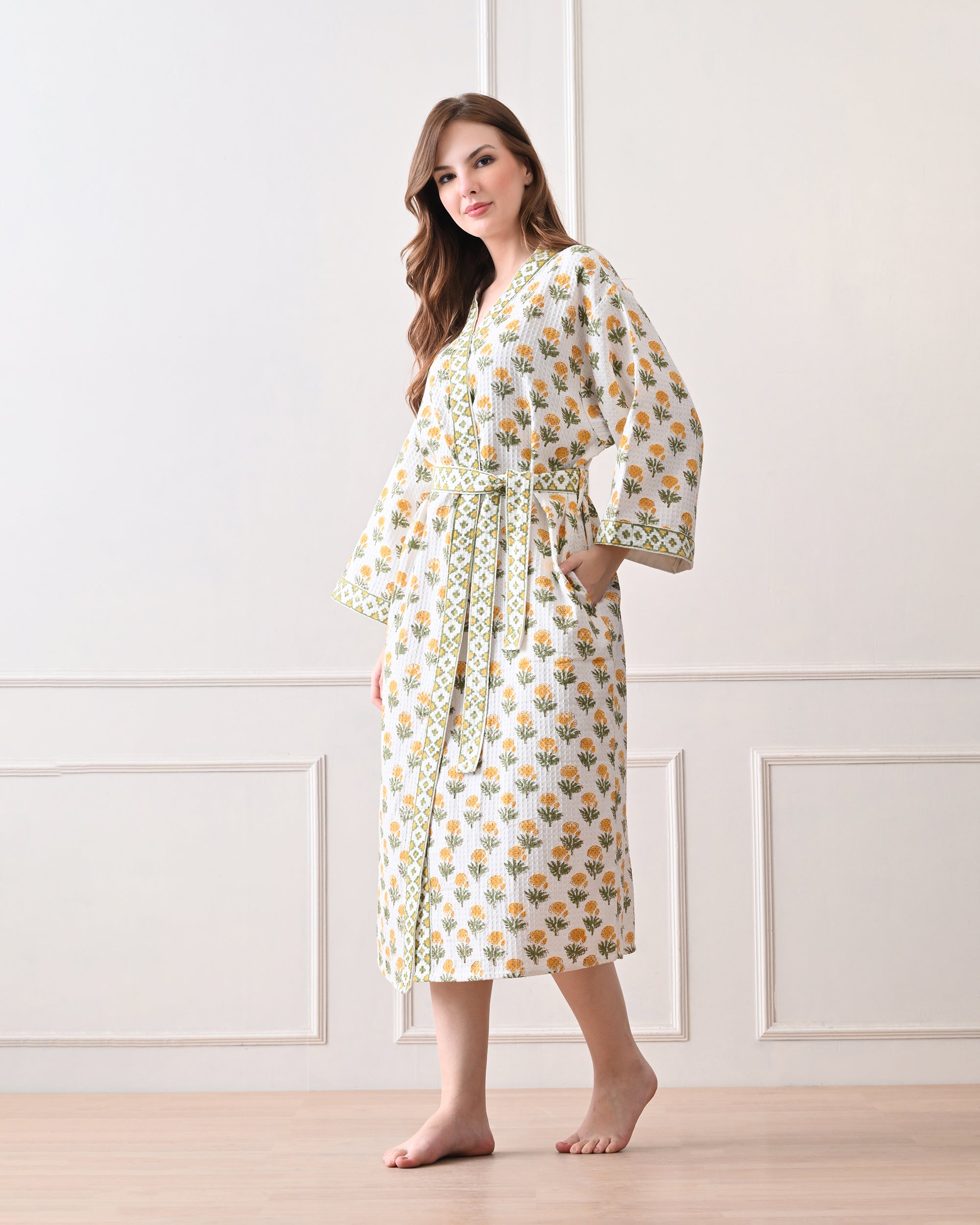 Sunrise Bloom Yellow Cotton Waffle Bathrobe with hand block printed yellow and orange flowers with green stems, green and white geometric border, long sleeves, and waist tie belt, worn by a woman | Singhvis
