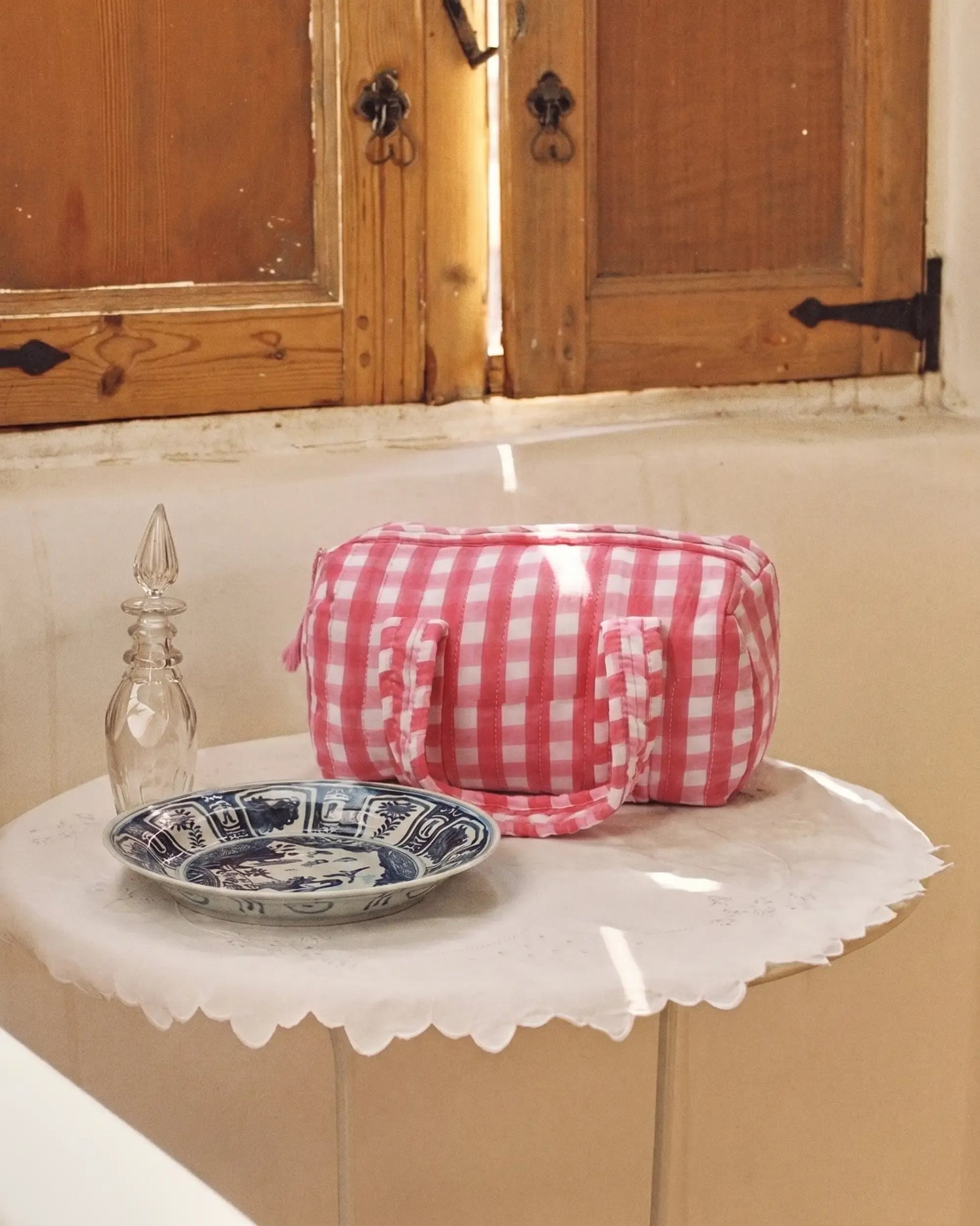 Pink gingham vanity bag with quilted texture and soft handles placed on a small table with a cloth, next to a glass bottle and a patterned bowl, with wooden window shutters in the background | Singhvis