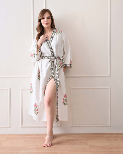 Botanic Whisper White Cotton Waffle Bathrobe with hand block printed pink tulip &amp; lotus-inspired floral patterns, green floral border, long sleeves, and waist tie belt, worn by a woman | Singhvis