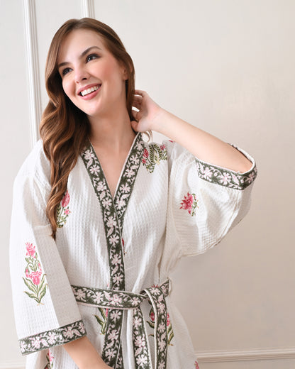 Botanic Whisper White Cotton Waffle Bathrobe with hand block printed pink tulip &amp; lotus-inspired floral patterns, green floral border, long sleeves, and waist tie belt, worn by a woman | Singhvis 