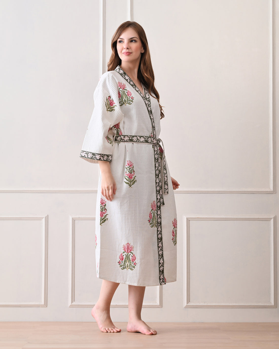 Botanic Whisper White Cotton Waffle Bathrobe with hand block printed pink tulip &amp; lotus-inspired floral patterns, green floral border, long sleeves, and waist tie belt, worn by a woman | Singhvis 