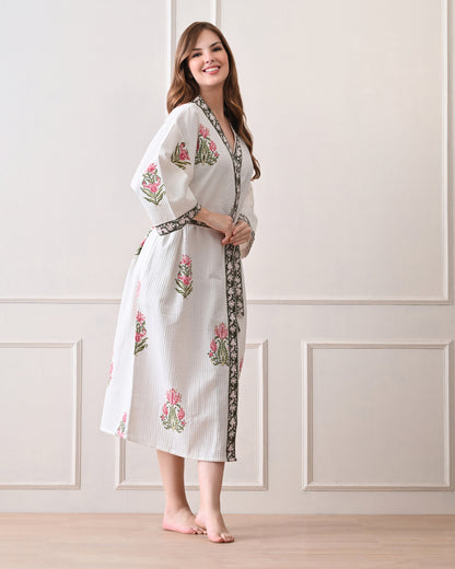 Botanic Whisper White Cotton Waffle Bathrobe with hand block printed pink tulip &amp; lotus-inspired floral patterns, green floral border, long sleeves, and waist tie belt, worn by a woman | Singhvis 