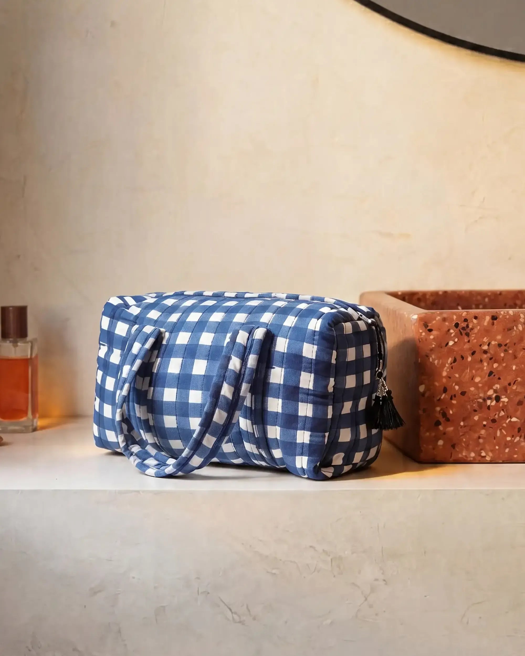 Blue gingham vanity bag with quilted texture and soft handles placed on a bathroom counter next to a bottle and a sink | Singhvis