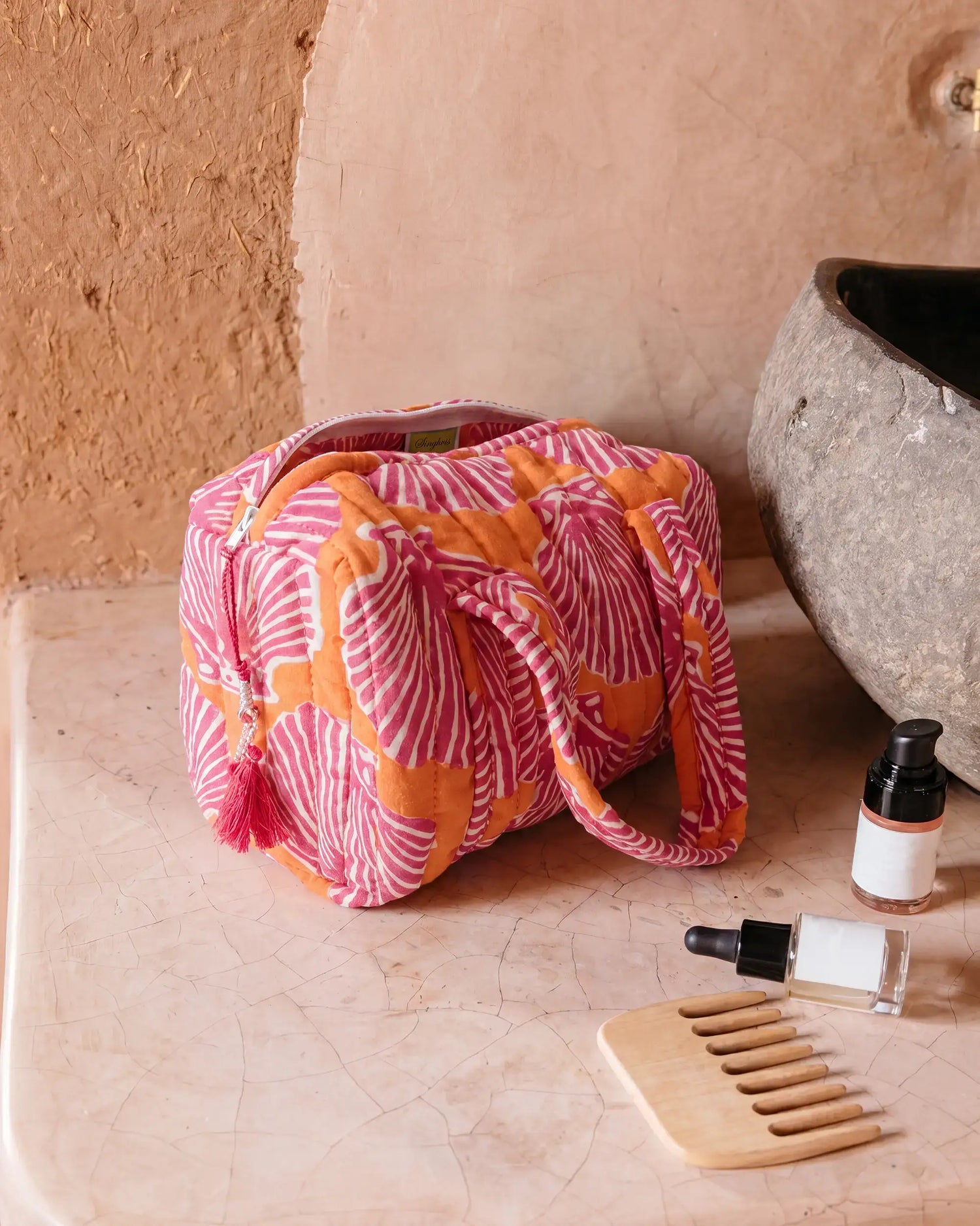 Orange and pink vanity bag with shell print and quilted texture on a bathroom counter, next to a sink, with a comb and small bottles placed nearby | Singhvis