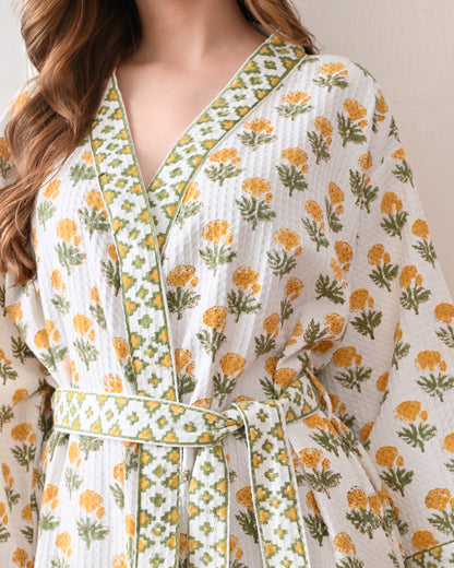 Sunrise Bloom Yellow Cotton Waffle Bathrobe with hand block printed yellow and orange flowers with green stems, green and white geometric border, long sleeves, and waist tie belt, worn by a woman | Singhvis